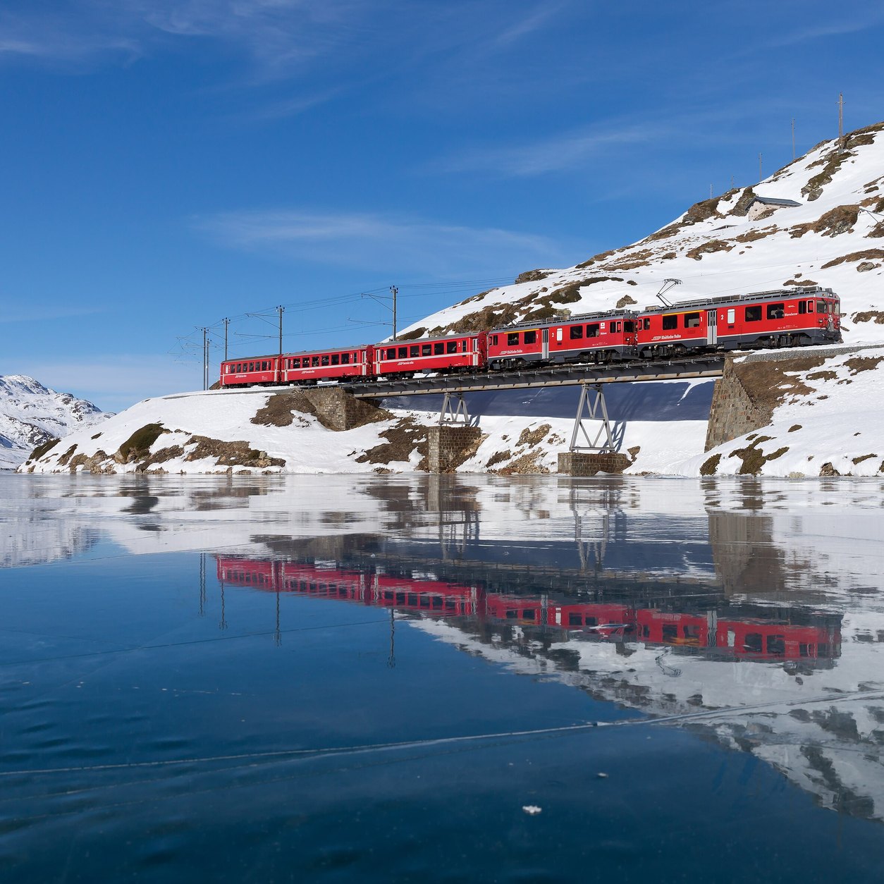 Et rødt tog fra Berninabanen krysser en bro ved Lago Bianco og speiler seg i vannet, med snø i fjellsiden